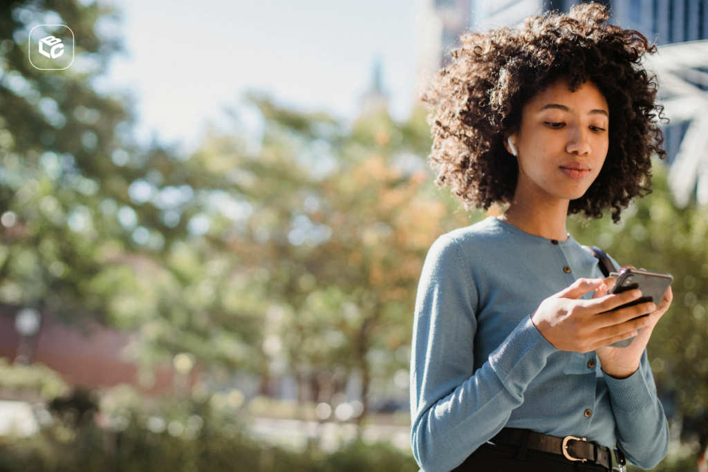 Mulher jovem com cabelo afro usando celular com fones de ouvido ao ar livre.