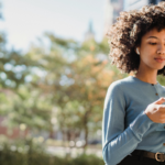 Mulher jovem com cabelo afro usando celular com fones de ouvido ao ar livre.