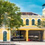 Edifício amarelo histórico com torre, em frente a uma árvore verde e fonte
